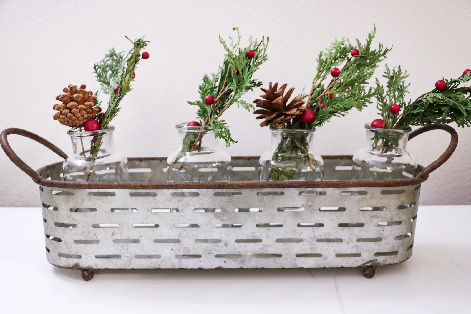 A galvanised trough centrepiece holding small glass jars of fresh greenery, red berries and pinecones