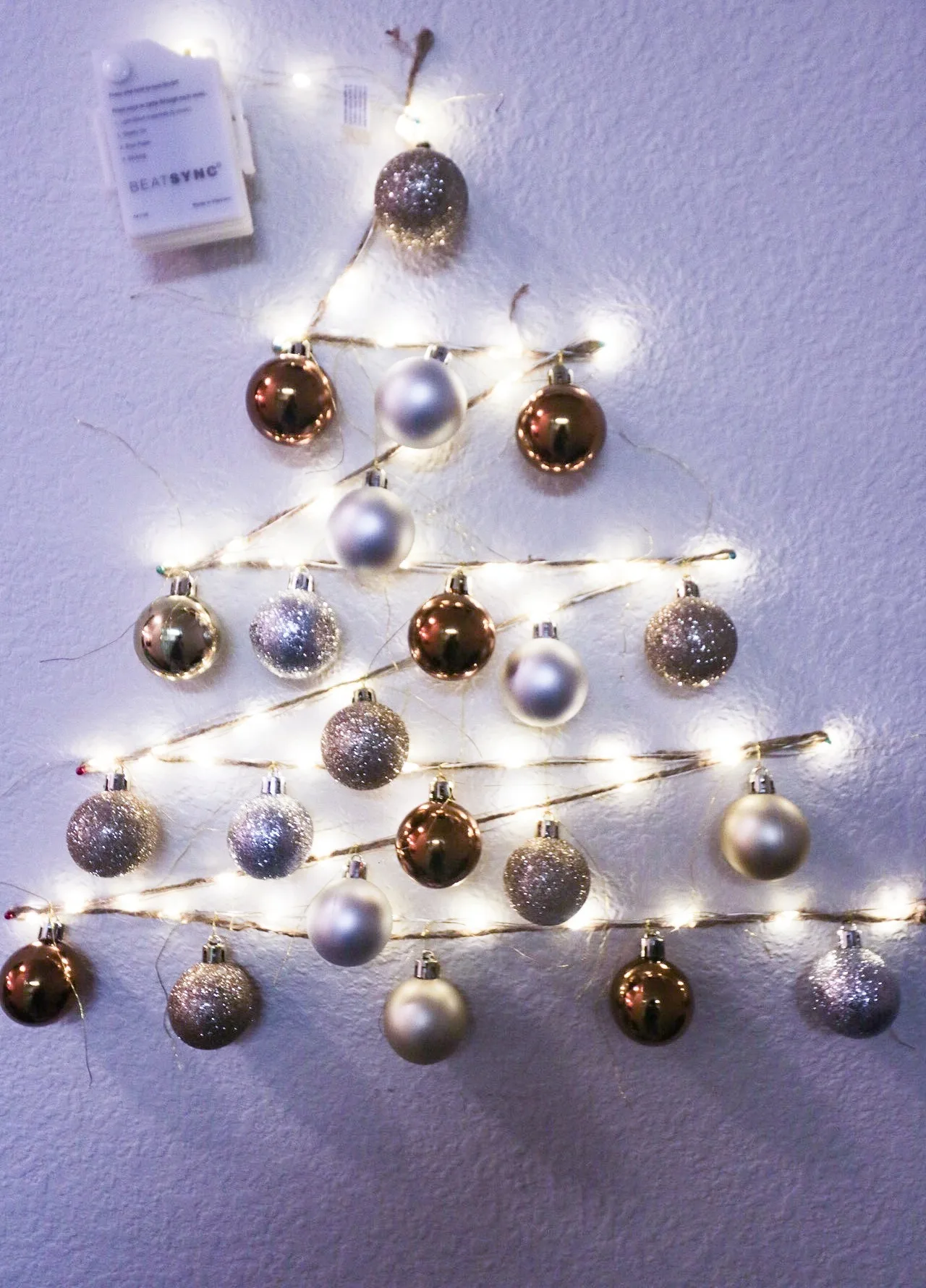 A Christmas tree shape made from fairy lights and baubles hung flat against a wall