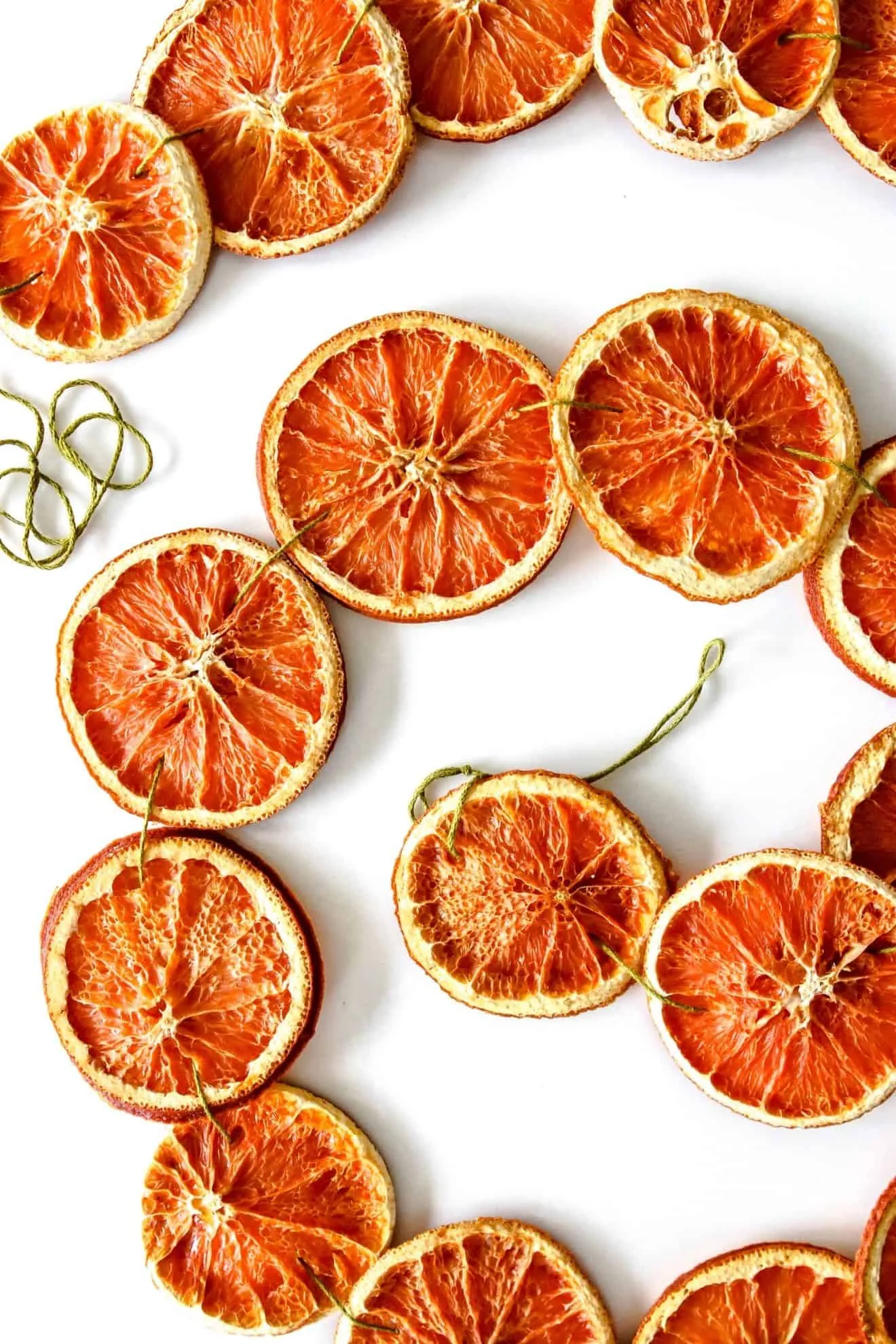 Dried orange slices laid out in a wavy line on white, each threaded with green twine, ready to be strung into a garland