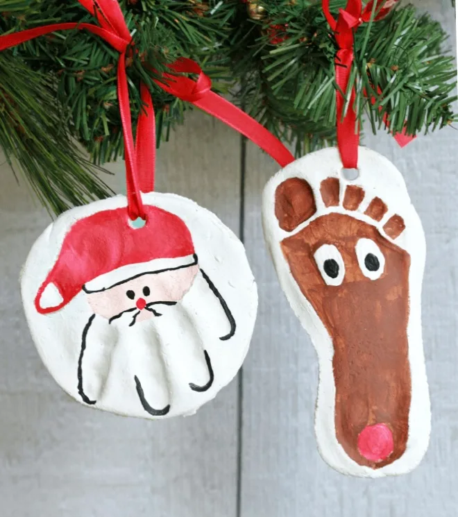 Two salt dough ornaments hanging from an evergreen branch on red ribbon — a handprint painted as Santa and a footprint painted as Rudolph